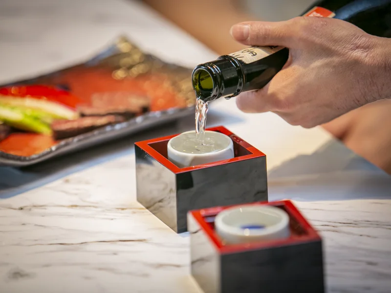 Sake being poured into a wooden masu cup at the marble counter